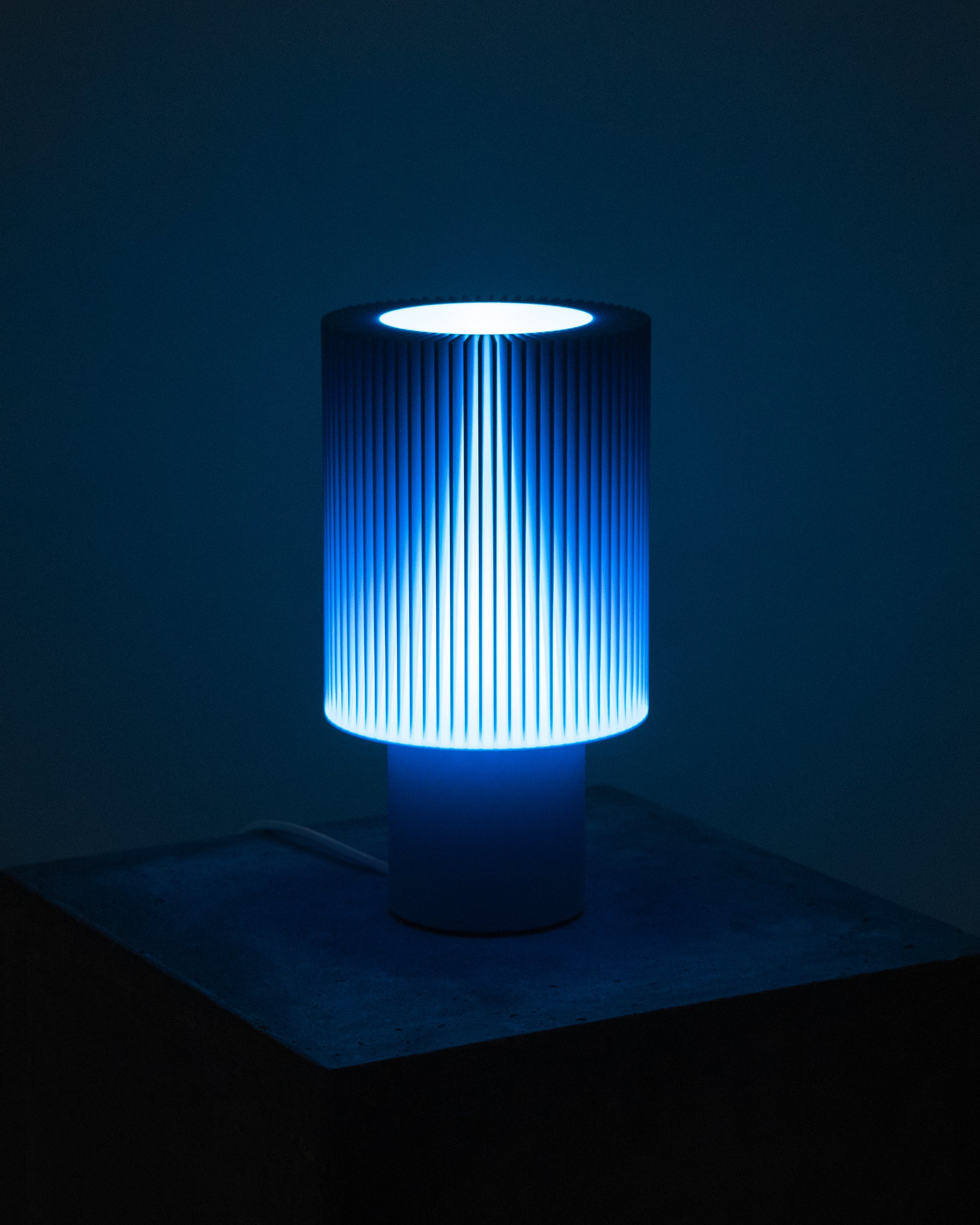 Lampe Iris Colors – Azur allumée, diffusant une lumière froide et électrique à travers un abat-jour nervuré, ambiance nocturne et contemporaine.